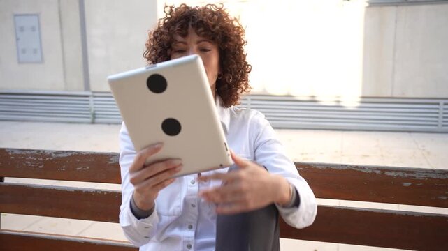 A young woman is using a tablet while sitting comfortably on a bench in an urban setting