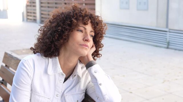 A thoughtful contemplative woman with curly hair sits in a peaceful urban setting, reflecting