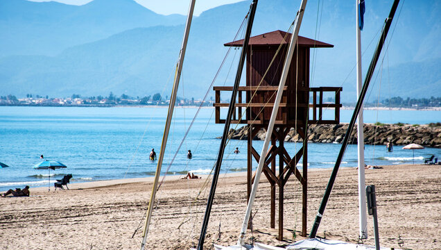 Torre de vigilancia de madera sobre la arena, junto a palmeras y m&aacute;stiles con bandera azul, s&iacute;mbolo de playa certificada. 