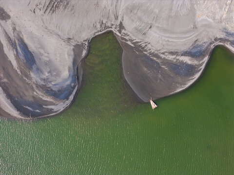 Aerial view of Depok Beach featuring dark volcanic sand contrasting with vibrant green water and a small traditional boat anchored along the shoreline in Special Region of Yogyakarta, Indonesia.