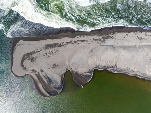 Aerial view of Depok Beach showing dark volcanic sand, crashing ocean waves, and the calm green waters of the lagoon in the Special Region of Yogyakarta, Indonesia.