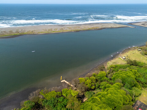 Aerial view of Depok Beach where the river meets the ocean, featuring a wooden pier, lush green vegetation, and sandy coastline in Special Region of Yogyakarta, Indonesia.