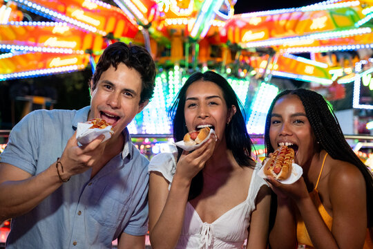 Three friends enjoying street food with bright amusement park lights in background