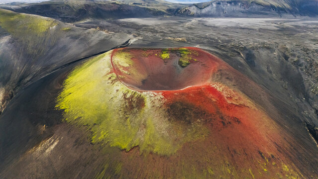 Aerial view of the vibrant red and green Raudaskal crater surrounded by dark volcanic landscape in Rangarthing ytra, Iceland.