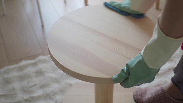 A person wearing protective gloves thoroughly cleans a small wooden round table, wiping its surface with a blue cloth and spray bottle