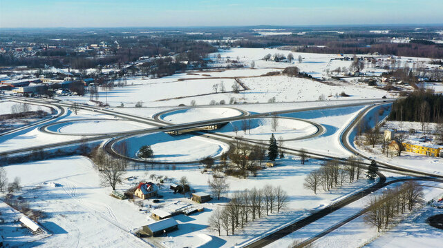 Aerial view of a cloverleaf interchange surrounded by snow-covered fields, trees, and rural buildings in Latvia.