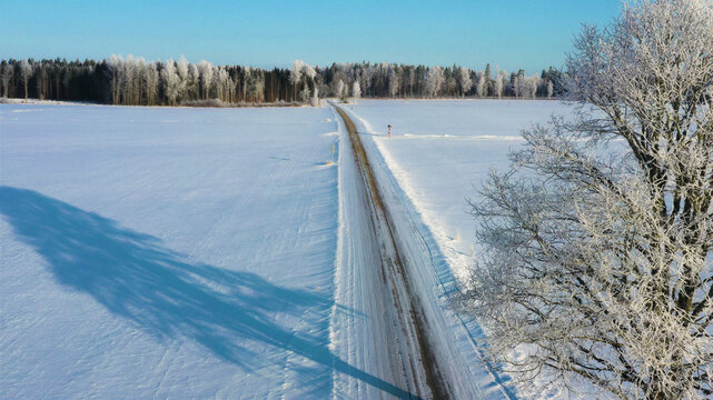 Aerial view of a straight road cutting through a vast snow-covered landscape with frost-covered trees under a clear blue sky in Latvia.