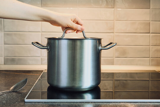 Hand lifting lid from stainless steel pot on modern induction cooktop, preparing food in kitchen
