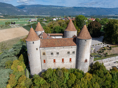 Aerial view of Champvent Castle with its distinctive round towers and terracotta roofs, surrounded by lush trees and rolling hills in Champvent, Vaud, Switzerland.