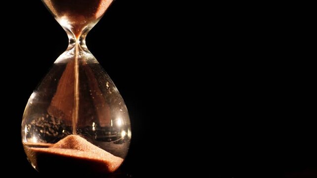 Close up of sand flowing through glass hourglass on black background representing time passing.