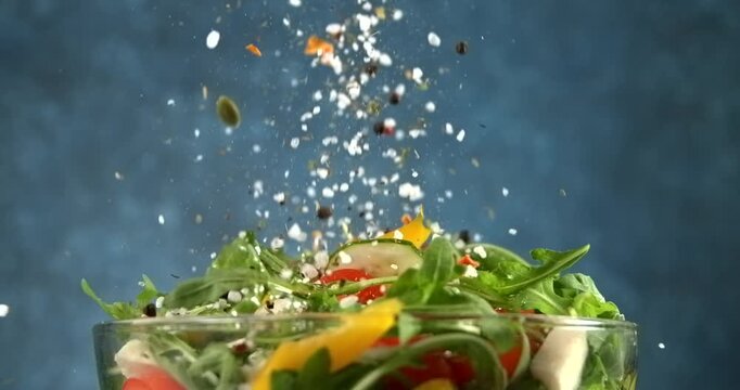 Herbs and spices falling onto fresh vegetable salad with arugula tomato cucumber and cheese in slow motion
