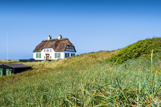 Traditional Danish thatched roof house standing on a grassy hill near the coast in Hirtshals, Denmark. Scenic historical cottage building under a clear blue sky.