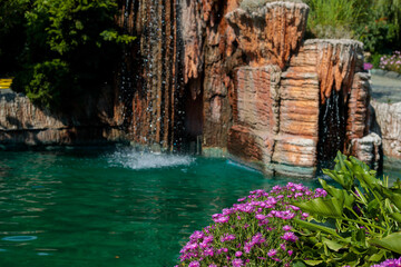 Artificial rock waterfall cascading over stone formations into turquoise pool surrounded by pink flowers, luxury resort landscape closeup with lush green foliage, high quality photo © Татьяна Швец