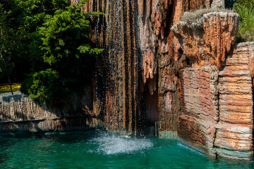 Artificial rock waterfall cascading over stone formations into turquoise pool surrounded by pink flowers, luxury resort landscape closeup with lush green foliage, high quality photo © Татьяна Швец