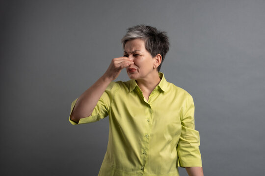 Senior woman pinching her nose because of bad odor and stinky smell in grey studio