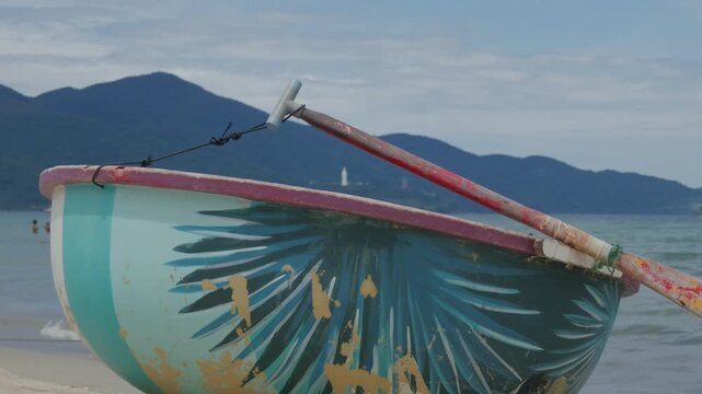 Painted wooden rowboat bow with lashed oar resting on sandy beach, turquoise and teal hull paint, chipped red paddle grip, overcast coastal sky