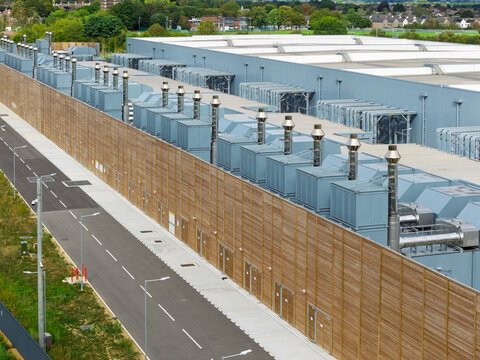Aerial view of a large industrial data center facility featuring rows of cooling units, a long wooden perimeter fence, and an adjacent road, United Kingdom.