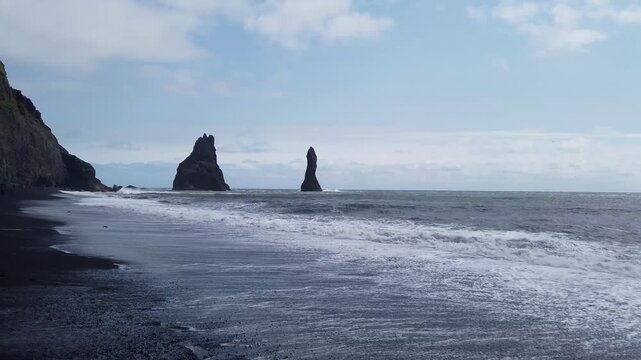 Reynisdrangar sea stacks rising offshore from Reynisfjara black sand beach near Vik, Iceland, with Atlantic waves washing over the shore under a blue sky.