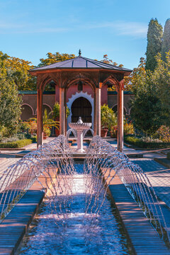 A symmetrical water channel with arcing jets leads toward a carved pavilion and arched doorway in a richly landscaped Islamic inspired garden.