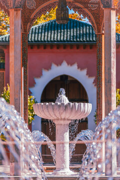 A white marble fountain stands between sparkling water jets beneath carved arches, with a terracotta wall and dark doorway beyond.
