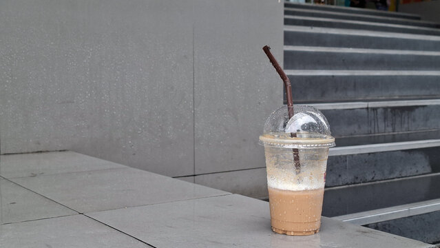 Disposable plastic cup of iced coffee with straw placed on outdoor concrete stairs ledge in urban building area