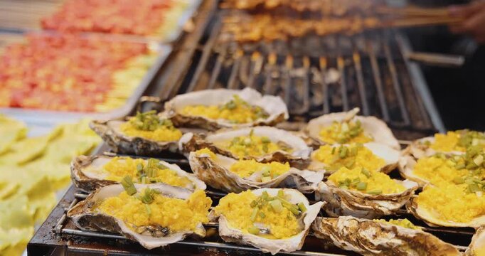 Fresh oysters topped with minced garlic and green onions roasting on a grill at a night market, traditional Asian street food preparation, culinary travel documentary.