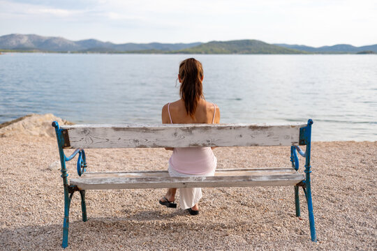 A long-haired brunette sits alone on a wooden bench overlooking the sea. She gazes peacefully toward the horizon, surrounded by a calm coastal landscape and soft natural light. 