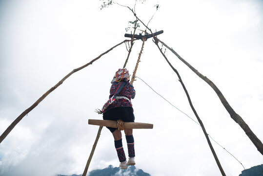 Young Akha hill tribe girl in vibrant traditional Akha costume sitting on wooden swing surrounded by lush green forest in Northern Thailand..