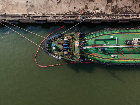 Aerial view of a tugboat moored at the Port of Tanjung Intan Cilacap with a green deck and oil containment boom alongside the concrete pier in Cilacap, Central Java, Indonesia.