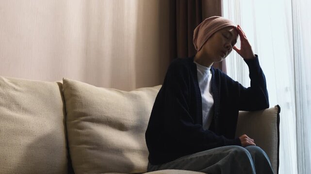 Asian woman wearing a headscarf sitting indoors with head in hand suffering from headache. Cancer patient, chemotherapy side effect, stress, and healthcare.
