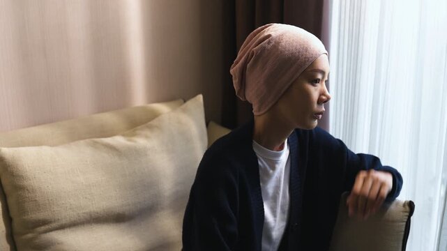 Asian woman wearing a headscarf sitting indoors with head in hand suffering from headache. Cancer patient, chemotherapy side effect, stress, and healthcare.