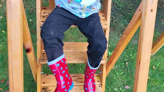 Child wearing red rain boots and black pants climbing wooden outdoor steps in a garden.
