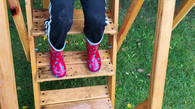 Child wearing red rain boots and black pants climbing wooden outdoor steps in a garden.
