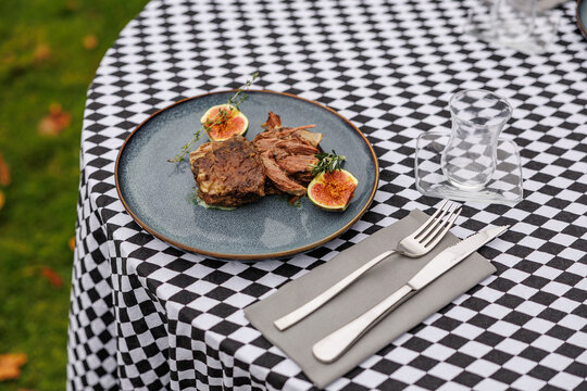 Cooked lamb pieces with fresh figs, rosemary, and thyme on a blue ceramic plate, set on a black-and-white checkered tablecloth with cutlery
