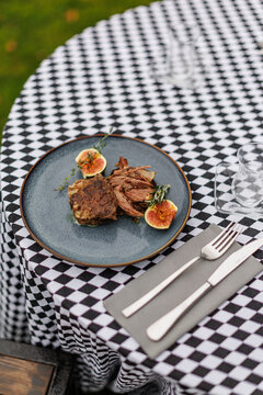 Cooked lamb pieces with fresh figs, rosemary, and thyme on a blue ceramic plate, set on a black-and-white checkered tablecloth with cutlery
