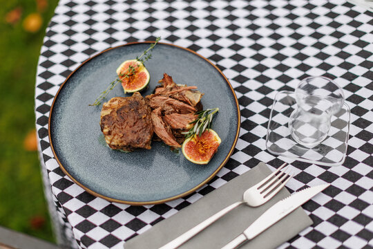Cooked lamb pieces with fresh figs, rosemary, and thyme on a blue ceramic plate, set on a black-and-white checkered tablecloth with cutlery