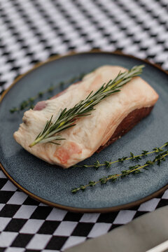 Raw lamb cut topped with fresh rosemary and thyme on a blue ceramic plate, accompanied by fork and knife on a black-and-white checkered tablecloth