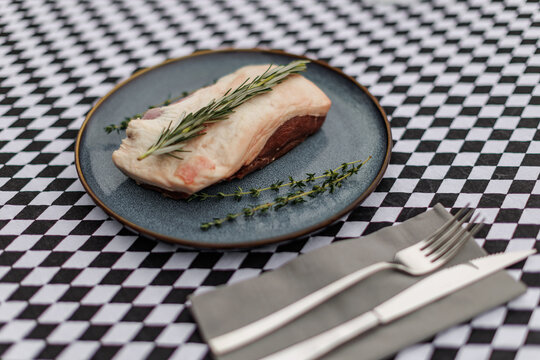 Raw lamb cut topped with fresh rosemary and thyme on a blue ceramic plate, accompanied by fork and knife on a black-and-white checkered tablecloth