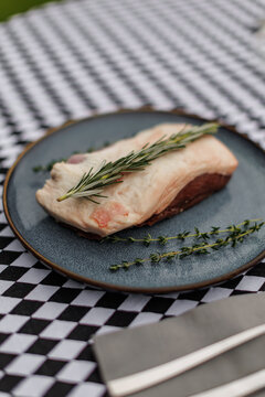 Raw lamb cut topped with fresh rosemary and thyme on a blue ceramic plate, accompanied by fork and knife on a black-and-white checkered tablecloth