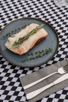 Raw lamb cut topped with fresh rosemary and thyme on a blue ceramic plate, accompanied by fork and knife on a black-and-white checkered tablecloth