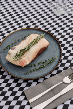 Raw lamb cut topped with fresh rosemary and thyme on a blue ceramic plate, accompanied by fork and knife on a black-and-white checkered tablecloth