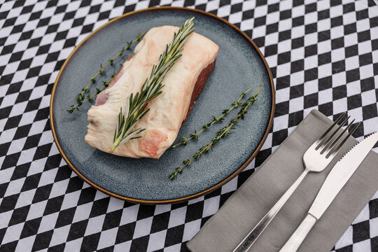Raw lamb cut topped with fresh rosemary and thyme on a blue ceramic plate, accompanied by fork and knife on a black-and-white checkered tablecloth