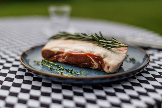 Raw lamb cut on a dark ceramic plate, topped with rosemary and thyme on a black-and-white checkered tablecloth in an outdoor setting
