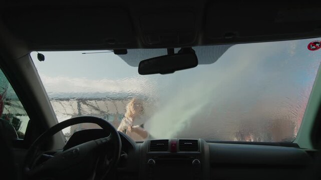 Woman washing car windshield with water pressure hose at outdoor self service station view from inside.