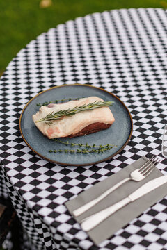 Raw lamb meat on a dark ceramic plate, garnished with fresh rosemary and thyme over a black-and-white checkered tablecloth