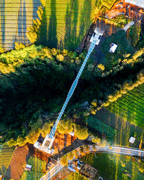 Aerial view of Jembatan Kaca glass bridge spanning a deep forested ravine surrounded by lush agricultural fields and terraced farmland at golden hour in Kecamatan Sukapura, Jawa Timur, Indonesia.