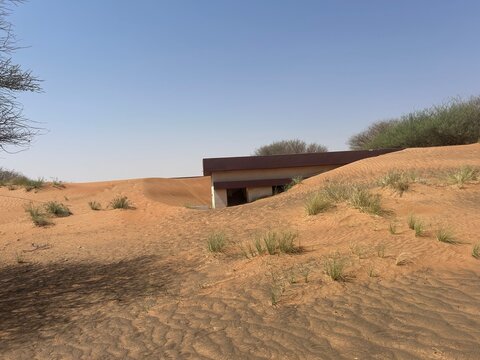 Abandoned house buried in desert sand dunes in United Arab Emirates landscape, showing nature reclaim human structure with dry vegetation