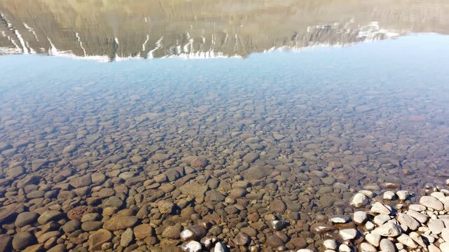 Lake Ljosavatn, Iceland, crystal clear shallow water over round pebbles, with a snow-streaked mountain reflected on the calm surface.