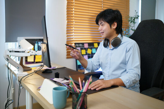 Young Asian male graphic designer holding stylus pen at creative workstation with drawing tablet, monitor, and sticky notes