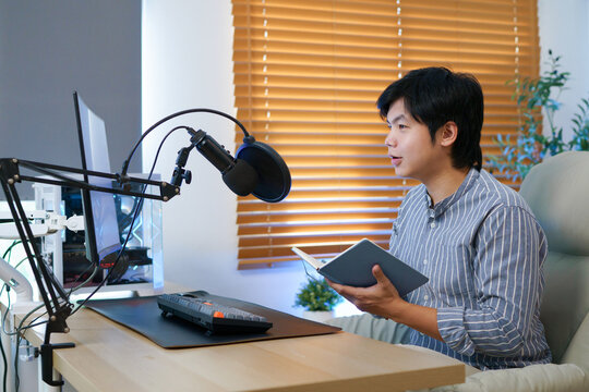 A young Asian male podcaster reads aloud from a notebook at his home recording desk, ideal for podcast, audiobook, and content creator concepts